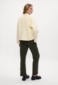 Kremowy sweter w splot warkoczowy, luźny krój, w połączeniu z ciemnozielonymi spodniami. Czarne buty na masywnej podeszwie. Minimalistyczne tło.