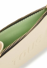 Bolso de color crema con forro interior verde, herrajes de cremallera dorados y detalle de logo grabado en el exterior.