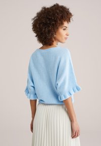 Pull en maille douce bleu clair avec manches bouffantes et poignets volantés, associé à une jupe plissée blanche. Vue de dos affichée.