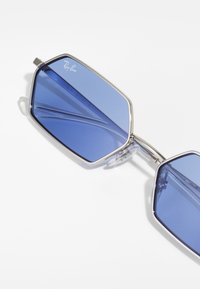 Ochelari de soare Hexagonali Ray-Ban au lentile de culoare albastră, cadre metalice și suporturi nazale transparente, evidențiind un design modern cu unghiuri ascuțite.