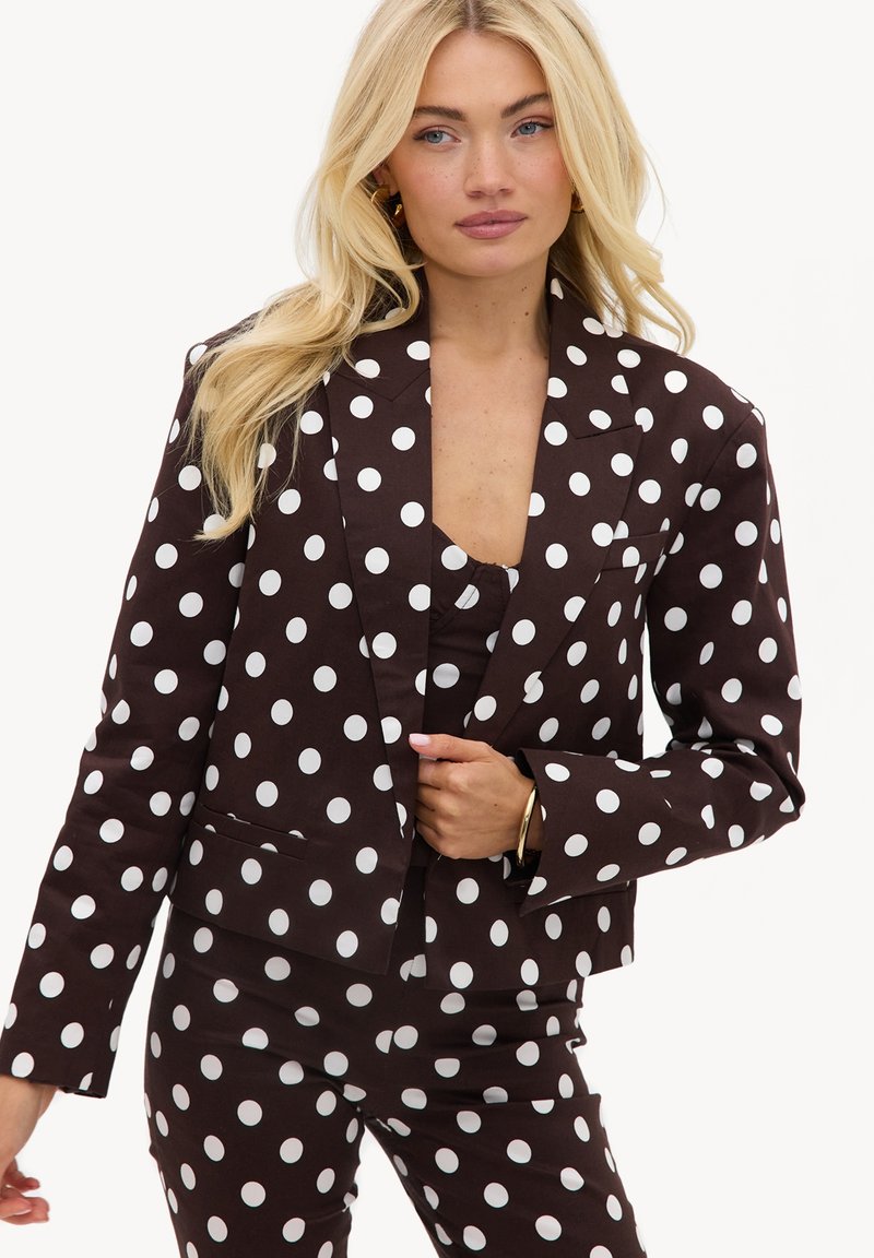 Femme blonde portant un costume noir à gros pois blancs, tenant le revers de la veste, sur fond blanc uni.