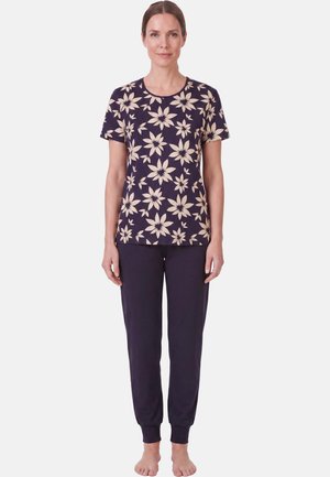 Vrouw staat op blote voeten, draagt een marineblauw shirt met korte mouwen en beige bloemenprint en een effen marineblauwe broek tegen een effen achtergrond.