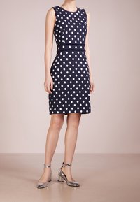 Námořnické modré šaty s puntíky, bez rukávů, s upnutým pasem a roztrhanými okraji. V kombinaci s kovově stříbrnými sandály na kotníky.