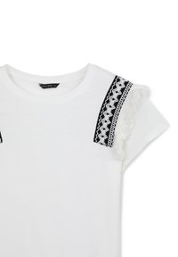 T-shirt bianco in cotone con maniche corte, dettagli in tessuto nero con motivi intricati e frange bianche sulle spalle.