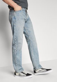 Vaaleansiniset farkut suoran lahkeiden leikkauksella, jossa on normaali vyötärö ja kontrastiväriset ompeleet. Yhdistetty mustiin ja valkoisiin lenkkareihin.