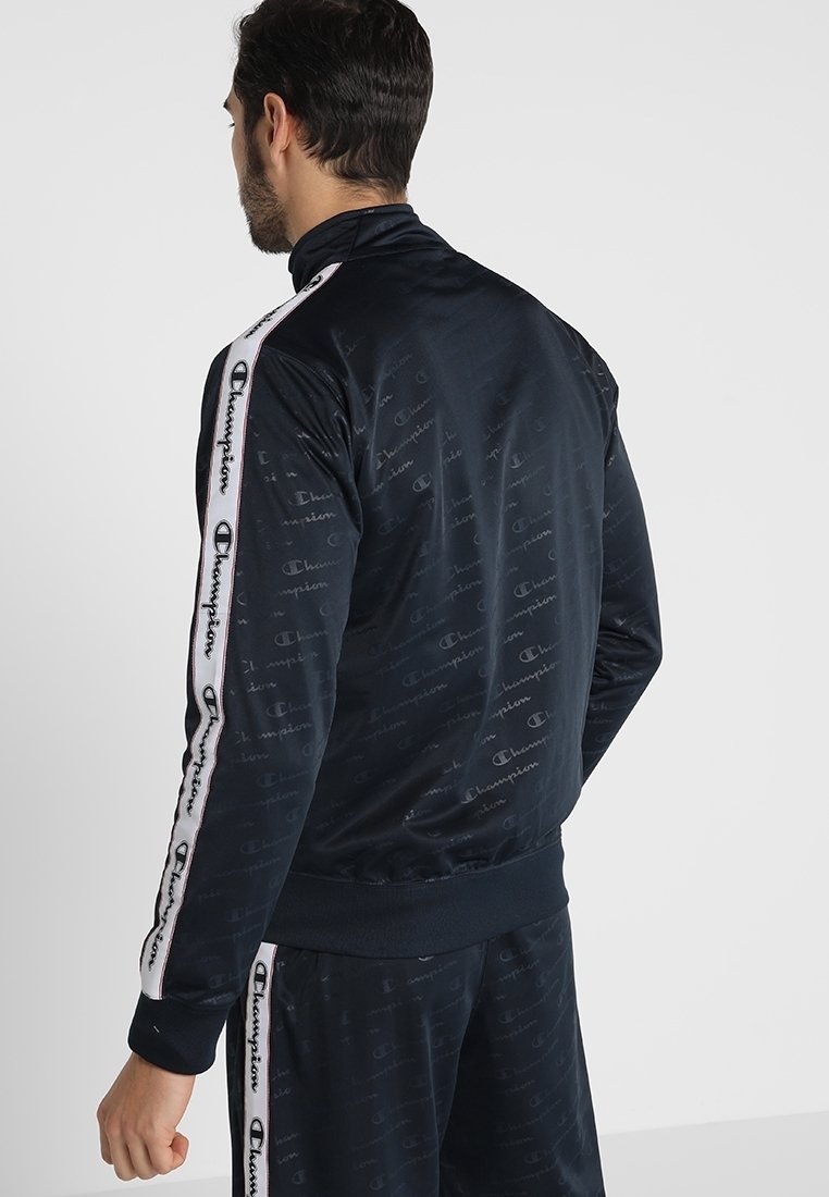 Marinblå träningsoveralljacka med hög krage, med ett upprepande logomönster, vita sidostråkar med varumärkeslogga och elastiska manschetter.