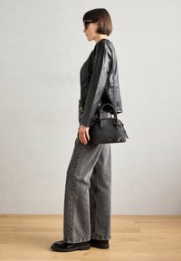 Borsa a mano in pelle nera con manici arrotondati, dettagli dorati e un fiocco. La modella indossa una giacca di pelle nera e pantaloni larghi grigi.