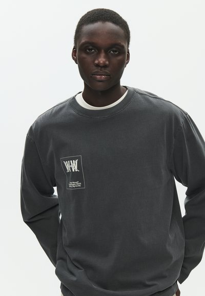 Jeune homme aux cheveux courts portant un sweat-shirt gris foncé avec un motif blanc imprimé sur le devant, debout devant un fond clair uni.