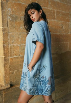 Frau mit langen dunklen Haaren, die ein hellblaues, kurzärmeliges Kleid mit Blumenmuster trägt und seitlich vor einer strukturierten Steinmauer steht.