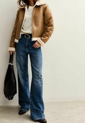 Une femme porte une veste beige doublée de peau lainée, un pull blanc, un jean large et des bottes. Elle tient un sac à main noir à franges.