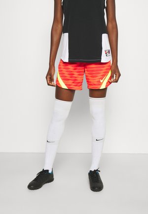 Person trägt ein schwarz-weißes Sporttop, rote gemusterte Shorts mit gelben Akzenten, weiße knielange Nike-Socken und schwarze Fußballschuhe.