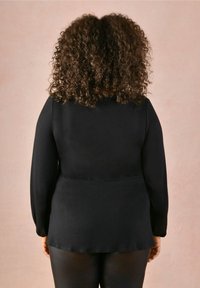 Långärmad svart topp med en slät textur och sammanknippade detaljer vid ärmarna, bärs över figursydda svarta leggings, sedd bakifrån.