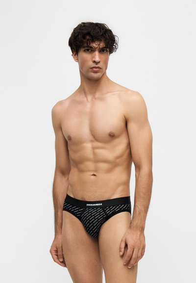 Homme musclé aux cheveux bouclés portant un slip noir à motifs avec le logo DSQUARED2 sur la ceinture, debout devant un fond clair uni.