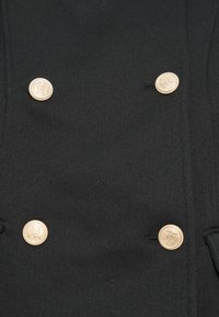 Μαύρο διπλής σειράς παλτό από υφασμένο ύφασμα, με τέσσερα κουμπιά σε χρυσή απόχρωση με ανάγλυφα σχέδια ως λεπτομερείς διακοσμήσεις.