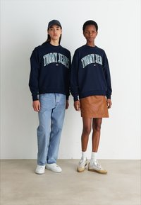 Sudadera azul marino con el texto "TOMMY JEANS", combinada con jeans de color azul claro y una falda de cuero marrón. Los accesorios incluyen una gorra y zapatillas blancas.