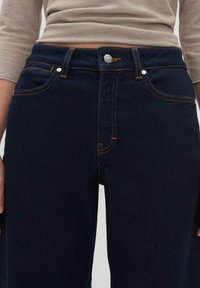 Jeans di denim blu scuro con cuciture arancioni prominenti, un classico design a cinque tasche e un bottone metallico argento in chiusura in vita.