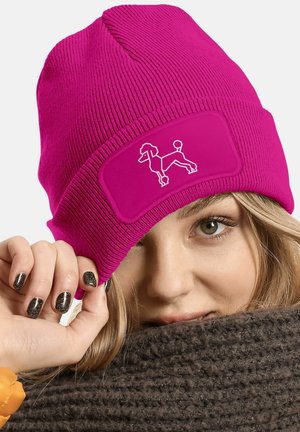 Fuchsia knit beanie with ribbed texture and a flat patch featuring a white poodle graphic. Worn partially over a brown scarf.