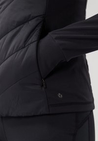 Chaqueta negra aislante con acabado liso, que cuenta con un bolsillo lateral con cremallera y una tela negra de textura contrastante en las mangas.