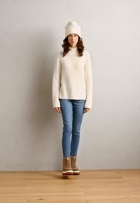 Kremfarget ribbestrikket genser, lyseblå skinny jeans og brune støvler med hvite såler. Modellen har på seg en hvit strikket lue; enkel bakgrunn.
