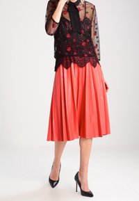 Blusa nera trasparente con ricami floreali e un fiocco, abbinata a una gonna corallo plissettata. Tacchi alti neri completano l'outfit.