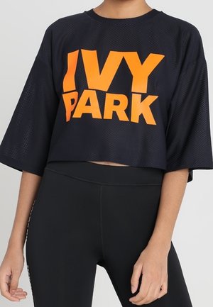 Kobieta w czarnej siateczkowej krótce skróconej bluzce z dużym pomarańczowym napisem "IVY PARK" oraz czarnych legginsach z wysokim stanem, stojąca na jednolitym tle.