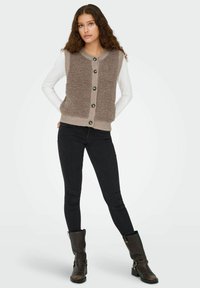 Braune ärmellose Weste mit Knöpfen aus strukturiertem Stoff, gerippten Kanten und grünen Knöpfen, kombiniert mit einem langärmeligen weißen Hemd und schwarzen Jeans.