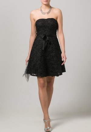 Femme portant une robe bustier en dentelle noire avec un nœud noué à la taille, un collier argenté, une pochette argentée et des chaussures à talons hauts argentées.