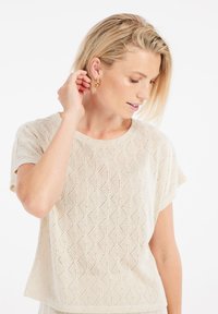 Maglia corta a maniche corte color beige chiaro con un motivo a rombi, caratterizzata da una vestibilità rilassata e un tessuto testurizzato. Orecchini a cerchio dorati completano il look.