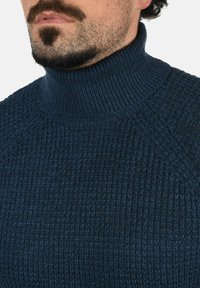 Jūras zilas krāsas augstais kakls svīteris ir ar strukturētu dizainu, kas raksturojas ar teksturētu adījumu un ribotiem akcentiem ap kaklu un pleciem.