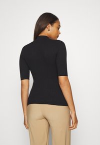 Black ribbed short-sleeve top with a high neckline, fitted silhouette, and textured fabric; paired with beige wide-leg pants.