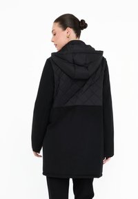 Černý kabát s prošívanou kapucí a horní částí zad, hladká tkanina a dlouhé rukávy. Design je minimalistický s volným střihem a bočními kapsami.