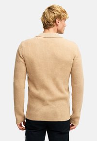 Beige strikket genser med strukturmønster, lange ermer og krage. Designet for en tettsittende passform med rett kant ved livet.
