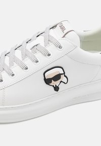 Vita lädersneakers med rund tå, texturerade grå snören och en plastkaraktärslogotyp på sidan. Texturerad gummisula.