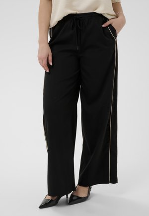 Kobieta ubrana w czarne szerokie spodnie z beżowym przeszyciem i czarne szpilki w szpic, stojąca z jedną ręką w kieszeni.