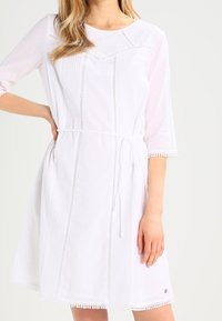 Robe blanche à manches trois-quarts, rayures verticales texturées, ceinture ajustée à la taille et bordure en dentelle le long des manches et de l'ourlet. Détail de logo circulaire à l'ourlet.