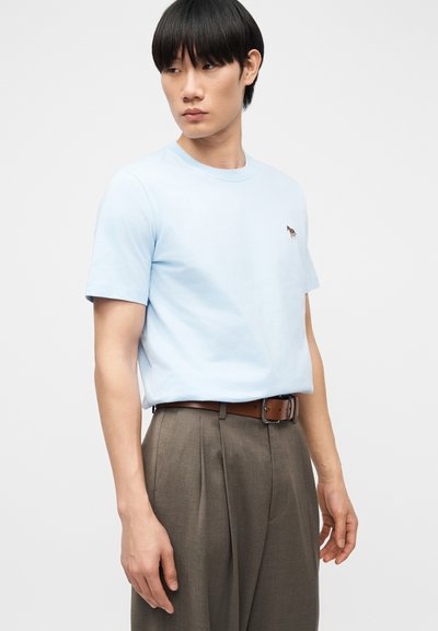 Νέος άνδρας με μαύρα μαλλιά που φοράει ανοιχτό γαλάζιο κοντομάνικο μπλουζάκι με ένα μικρό λογότυπο ζέβρας και καφέ ψηλόμεσα παντελόνια με ζώνη.