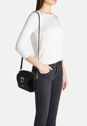 Donna che indossa una camicia bianca a maniche lunghe e jeans scuri con borsa a tracolla in pelle nera con accessori dorati e logo sul davanti.