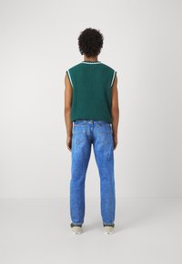 Un pulover verde tricotat fără mâneci, cu margini albe, asortat cu blugi drepți de culoare albastru deschis și adidași gri.