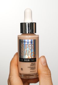 Maybelline Super Stay 24H Skin Tint w odcieniu 05, szklana butelka z kroplomierzem i srebrną nakrętką, gładka beżowa konsystencja, opatrzona szczegółową etykietą.
