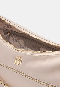Borsa a mano color beige chiaro con hardware dorato e dettaglio a catena. Presenta una chiusura con zip e un rivestimento in tessuto con logo e un motivo sottile.