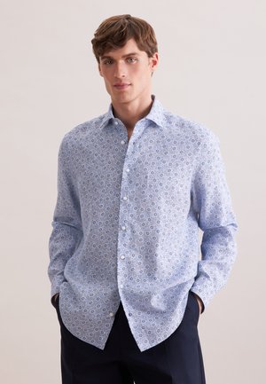 Młody mężczyzna w jasnoniebieskiej koszuli w wzory z rękami w kieszeniach ciemnych spodni, stojący na jednolitym tle.