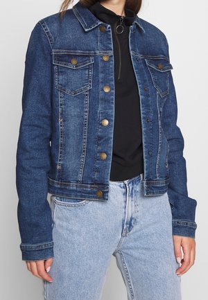 Kvinde iført en blå denimjakke med messingknapper over en sort lynlåshættetrøje og lyseblå jeans, står mod en ensfarvet baggrund.