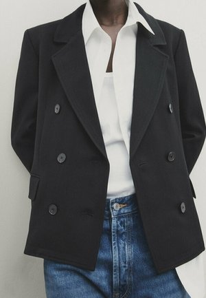 Personne portant un blazer noir croisé sur une chemise blanche rentrée dans un jean bleu taille haute.