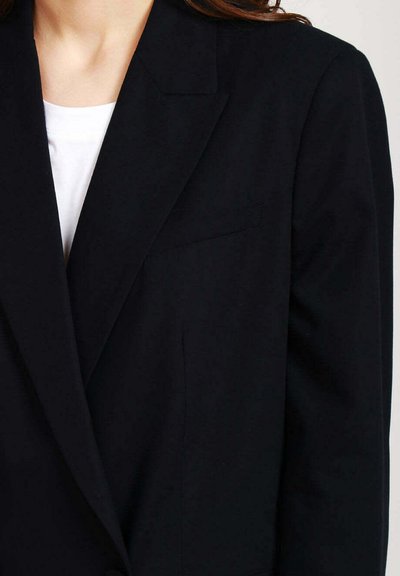 Blazer nero con rever a punta, taschino singolo e texture liscia. Indossato sopra una camicia bianca, mette in evidenza un design su misura.