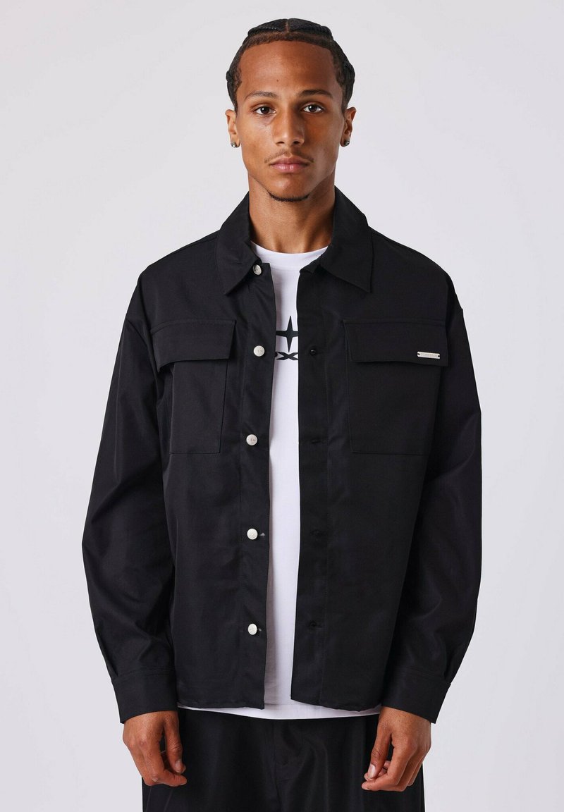 Giacca nera button-up realizzata con un materiale robusto, caratterizzata da quattro tasche frontali, bottoni a pressione bianchi e una leggera lucentezza. Indossata sopra una maglietta bianca.