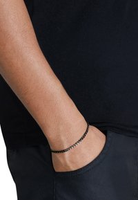 Bracciale in catena nera in metallo con anelli intrecciati, indossato su un polso che si estende da una tasca, caratterizzato da una superficie liscia e lucida.