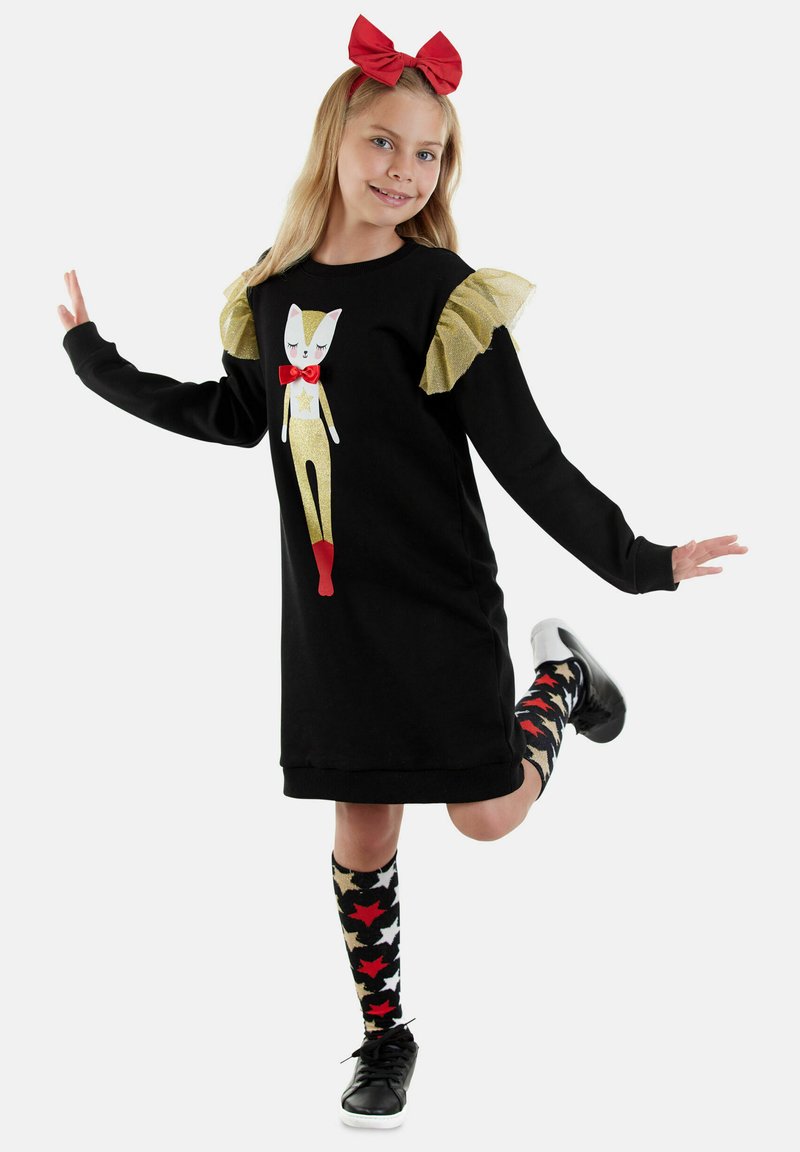 Vestido negro con mangas doradas con volantes, que presenta un gráfico de un gato con un lazo rojo y botas. Medias hasta la rodilla de rayas negro y rojo, zapatos negros.