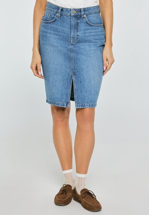 Kobieta ubrana w niebieską spódnicę dżinsową do kolan z przednim rozcięciem, w białych skarpetkach i brązowych mokasynach, stojąca z rozluźnionymi rękami.