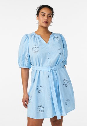 Kvinde iført en lyseblå, bælteklædt kjole med pufærmer og blomsterudskåret broderi, stående mod en enkel hvid baggrund.