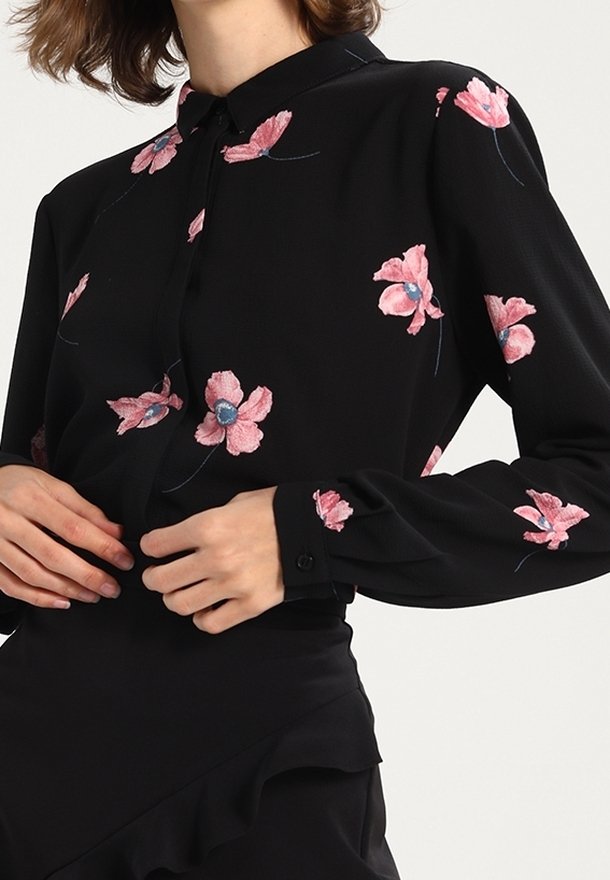Blouse noire à manches longues avec broderie florale rose portée par une personne ajustant le devant, associée à une jupe noire avec un détail à volants.